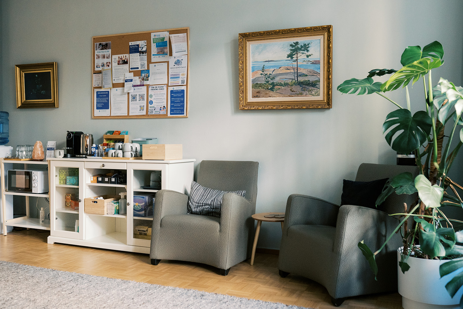 Compass Psychology Photo of waiting room with a shelf of teas and coffees, a notice board with therapy event advertsements, a framed scenery painting, and a pot plant in the corner.